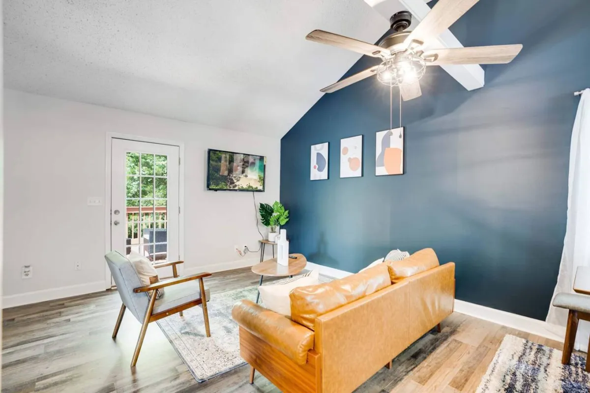 A stylish lounge area with a comfortable sofa, vibrant wall art, a television for entertainment, lush potted plant, and beautiful wooden floors. Sunlight filters through unseen windows, adding warmth to the space and hinting at the open, airy layout.