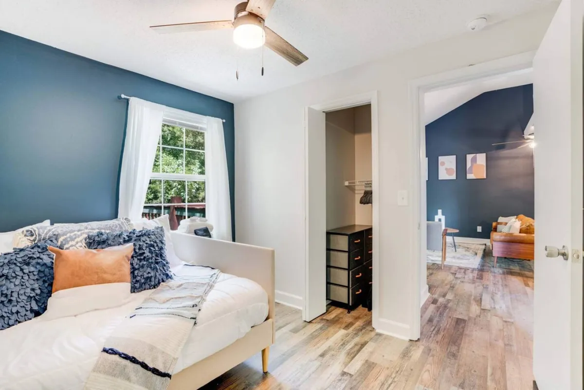 A cozy bedroom featuring a bed with decorative pillows, a window flooding the space with natural light, visible closet space, and an open doorway offering a peek into another room. The decor is soft and inviting, designed for comfort and relaxation.