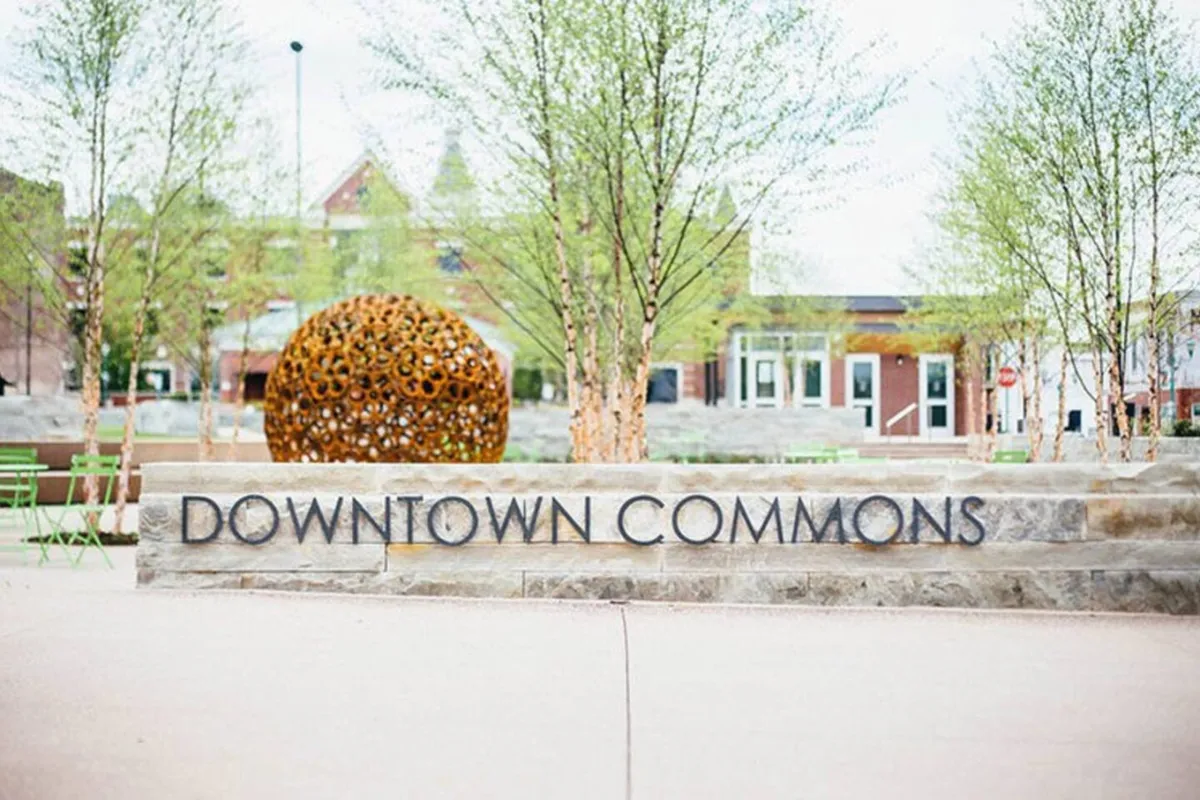 The scene showcases the Downtown Commons sign, an artistic outdoor sculpture, and leafy trees, highlighting a central gathering place. The mood is bright and welcoming, suggesting a hub for community events and cultural activities.