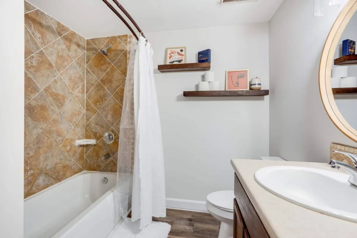 A modern bathroom with a sleek bathtub and shower area, circular mirror above a contemporary vanity, and shelves decorated with fresh touches. The space is bright, fresh, and designed for relaxation.