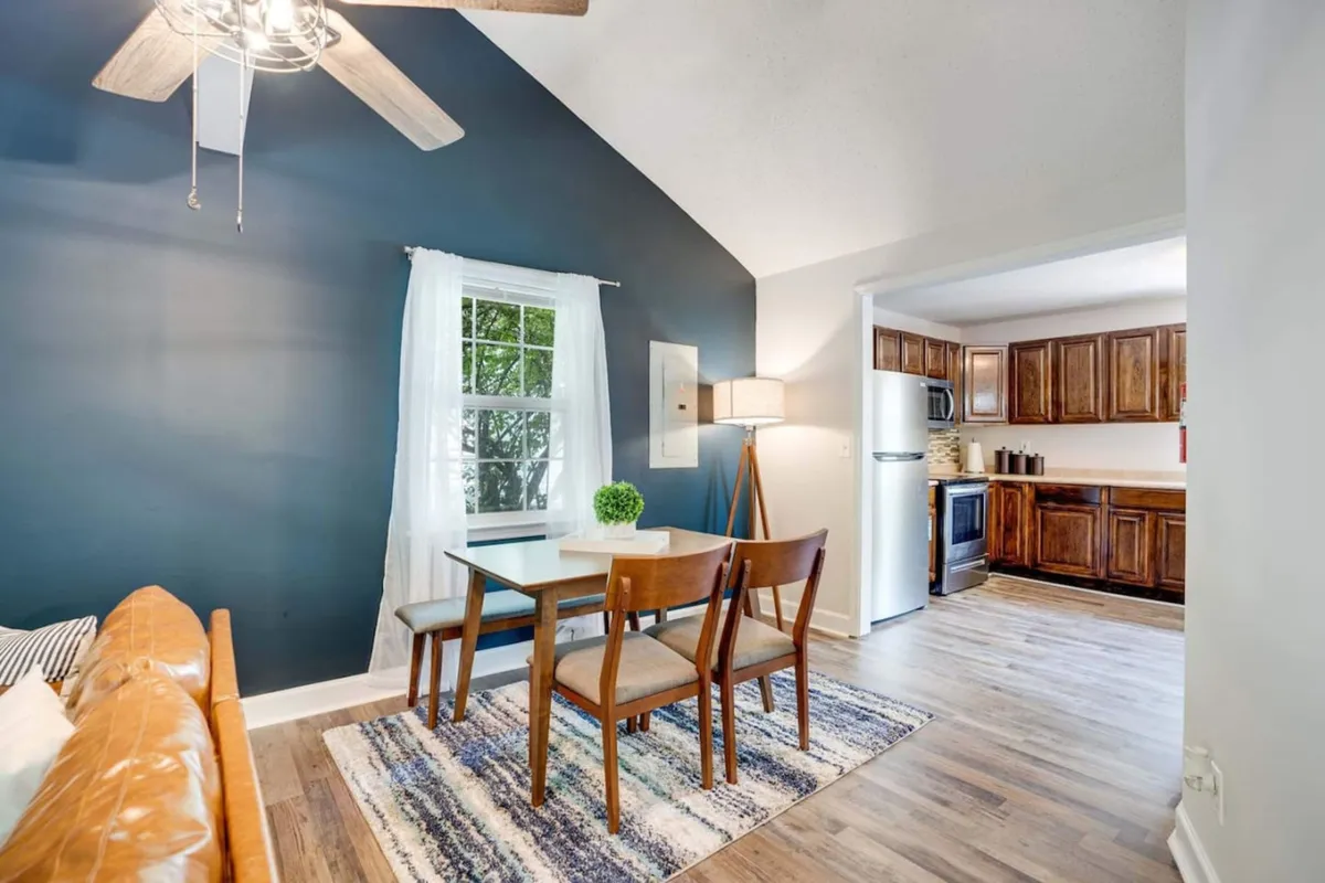 An open-concept living and dining area featuring a sofa, a well-set dining table, large window with curtains, and a glimpse of the adjoining kitchen. The scene exudes warmth and togetherness—ideal for family meals, casual gatherings, or quiet mornings.