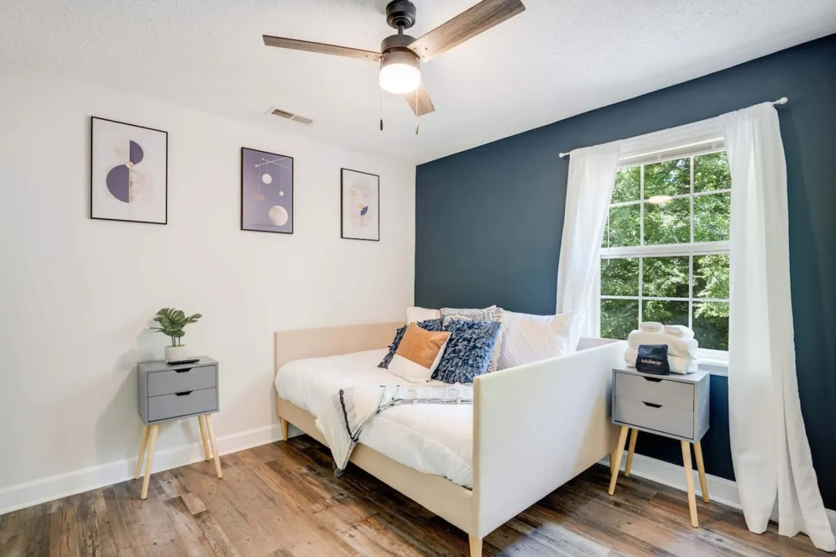 Another tranquil bedroom scene, with a bed adorned in plush pillows, natural light filtering through the window, and tasteful wall art that adds a serene touch. The overall mood is peaceful and conducive to relaxation.