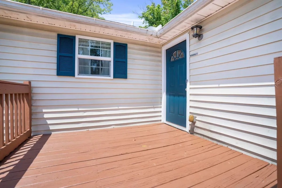 An inviting entrance view featuring a blue door, window shutters, and a wooden deck. The morning sunlight warms the deck, suggesting a pleasant transition from the comfort of home to the excitement of the city.