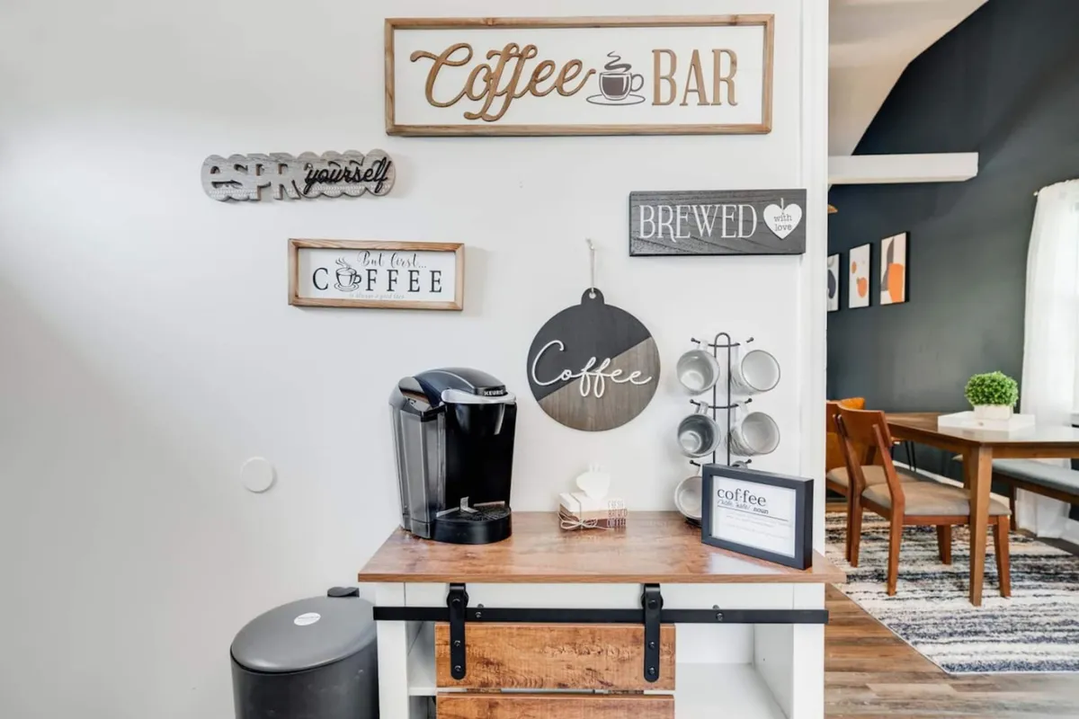 A cozy dining area and coffee bar setup with a coffee machine, decorative signs, and a welcoming ambiance. The space is designed for enjoying breakfast, brewing coffee, and socializing, with the dining area visible in the background.