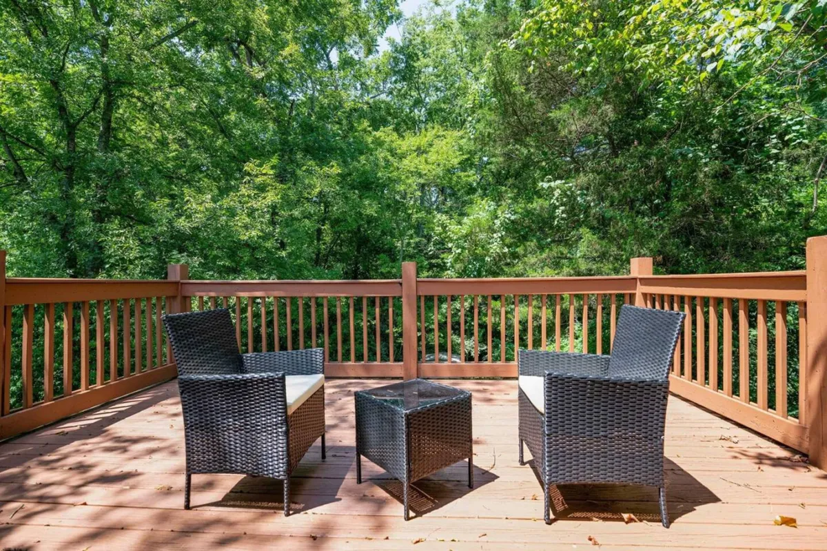 An inviting outdoor patio scene featuring wicker chairs gathered around a glass table, surrounded by a wooden railing and lush greenery beyond. The setting is tranquil, with soft natural light hinting at either morning or late afternoon, suggesting a pleasant space to enjoy the outdoors.
