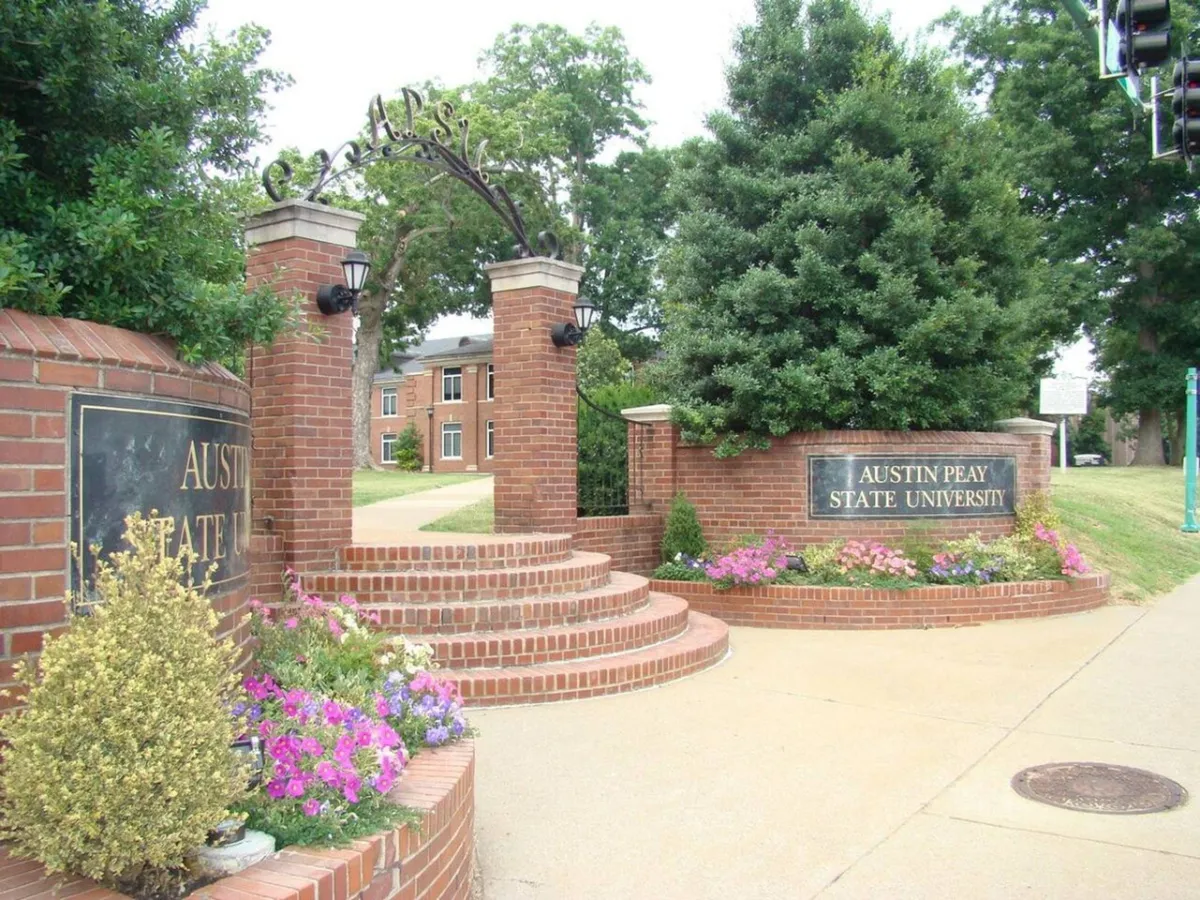 This image captures the brick entrance with clear university signage, surrounded by vibrant flower beds and well-maintained landscaping. The scene includes people walking and gathering, evoking a sense of arrival and campus life.