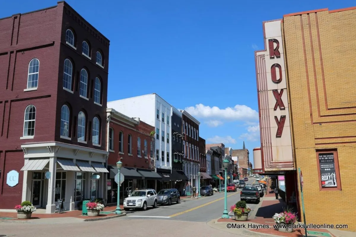 This exterior image captures the vibrant Roxy theatre signage, classic brick buildings lining the street, and cheerful flower pots. People can be seen strolling, emphasizing the lively atmosphere of downtown Clarksville.